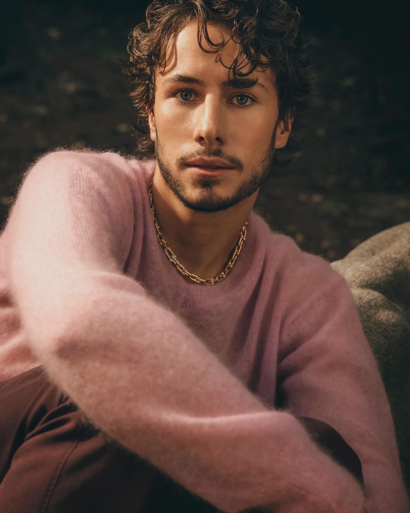 Close-up portrait of a man with curly dark hair wearing a pink fuzzy sweater and a gold chain necklace.