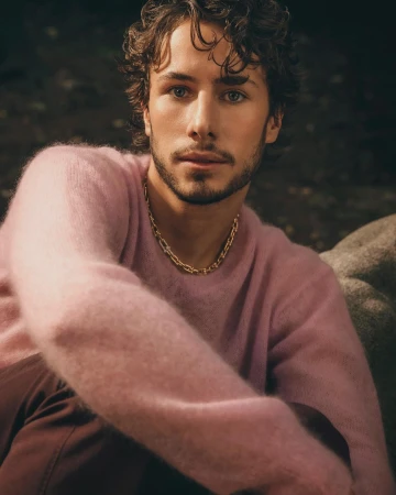 Close-up portrait of a man with curly dark hair wearing a pink fuzzy sweater and a gold chain necklace.