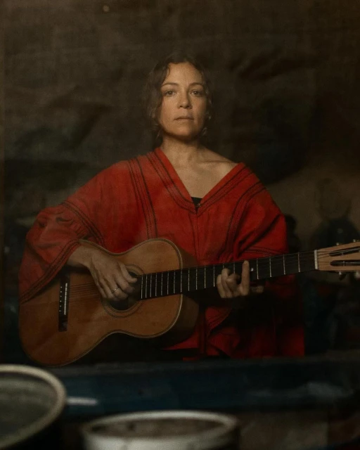 Woman in red top playing acoustic guitar indoors with vintage, muted colors. Portrait shot, looking directly at the camera.