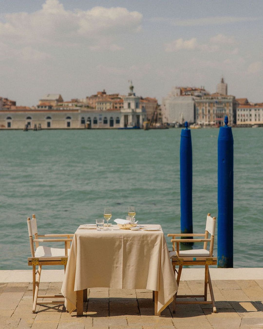 A table set for two with white linen and wine glasses, stands outdoors on a stone patio overlooking a scenic waterfront in Venice, Italy.