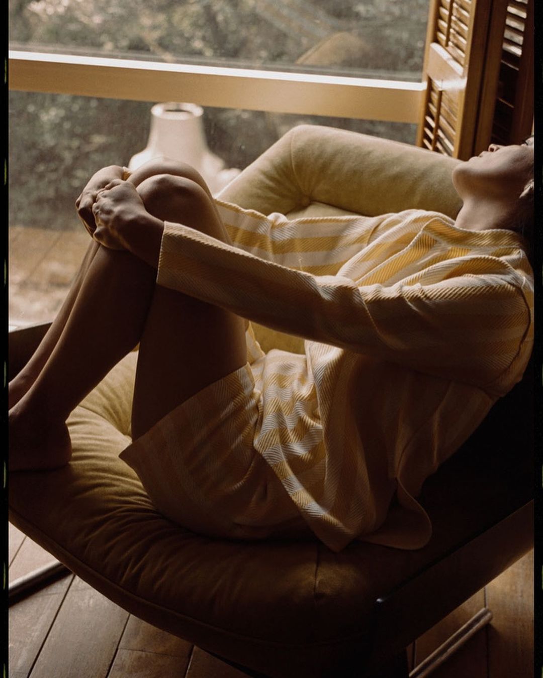Woman in yellow striped outfit lounges in an armchair by a sunlit window with wooden shutters, creating a warm and inviting scene.