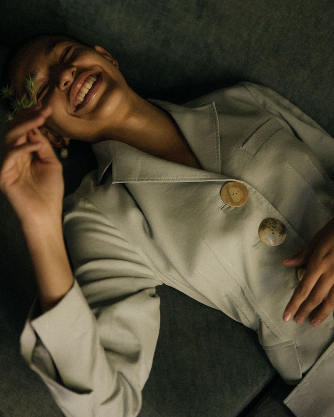 Smiling woman in an oversized gray blazer lying down with a small plant in her hand, lit with moody, diffused studio lighting.