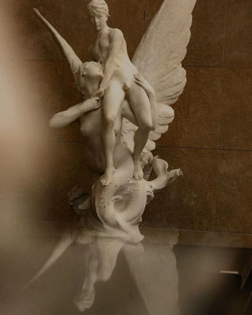 A white marble sculpture of a man with wings carrying a woman, with a reflection visible in the foreground.