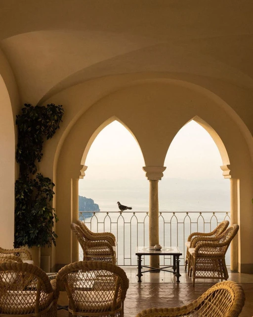 Wicker chairs arranged on a balcony overlooking the sea, viewed through arched windows in a naturally lit indoor space with a bird.