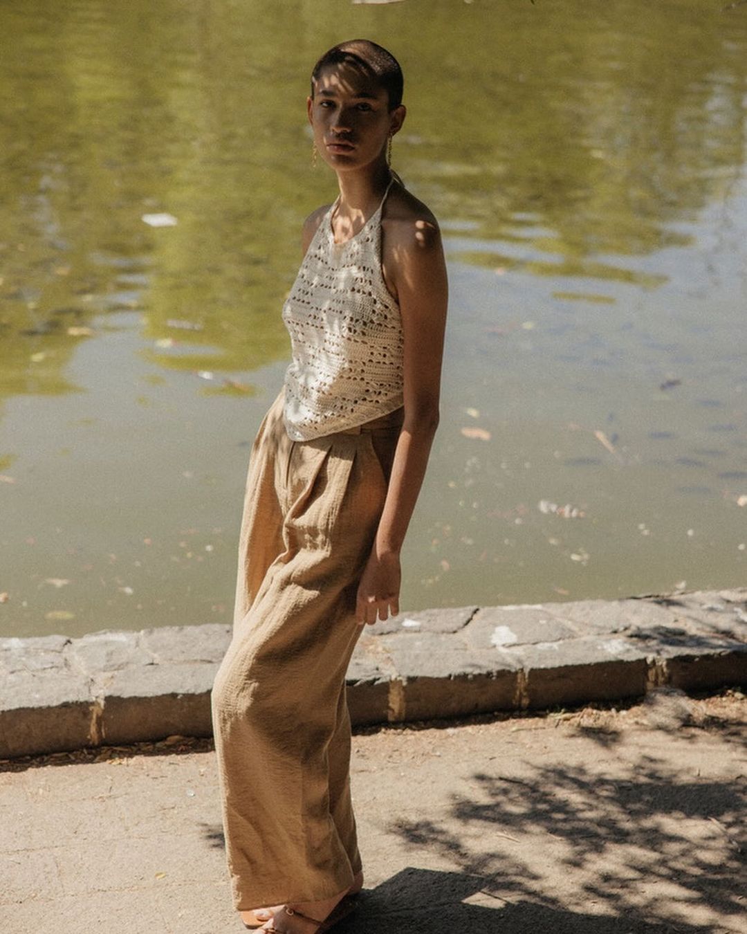 Woman in a crochet halter top and wide-leg pants poses outdoors with a lake in the background on a sunny day. Fashion shot.