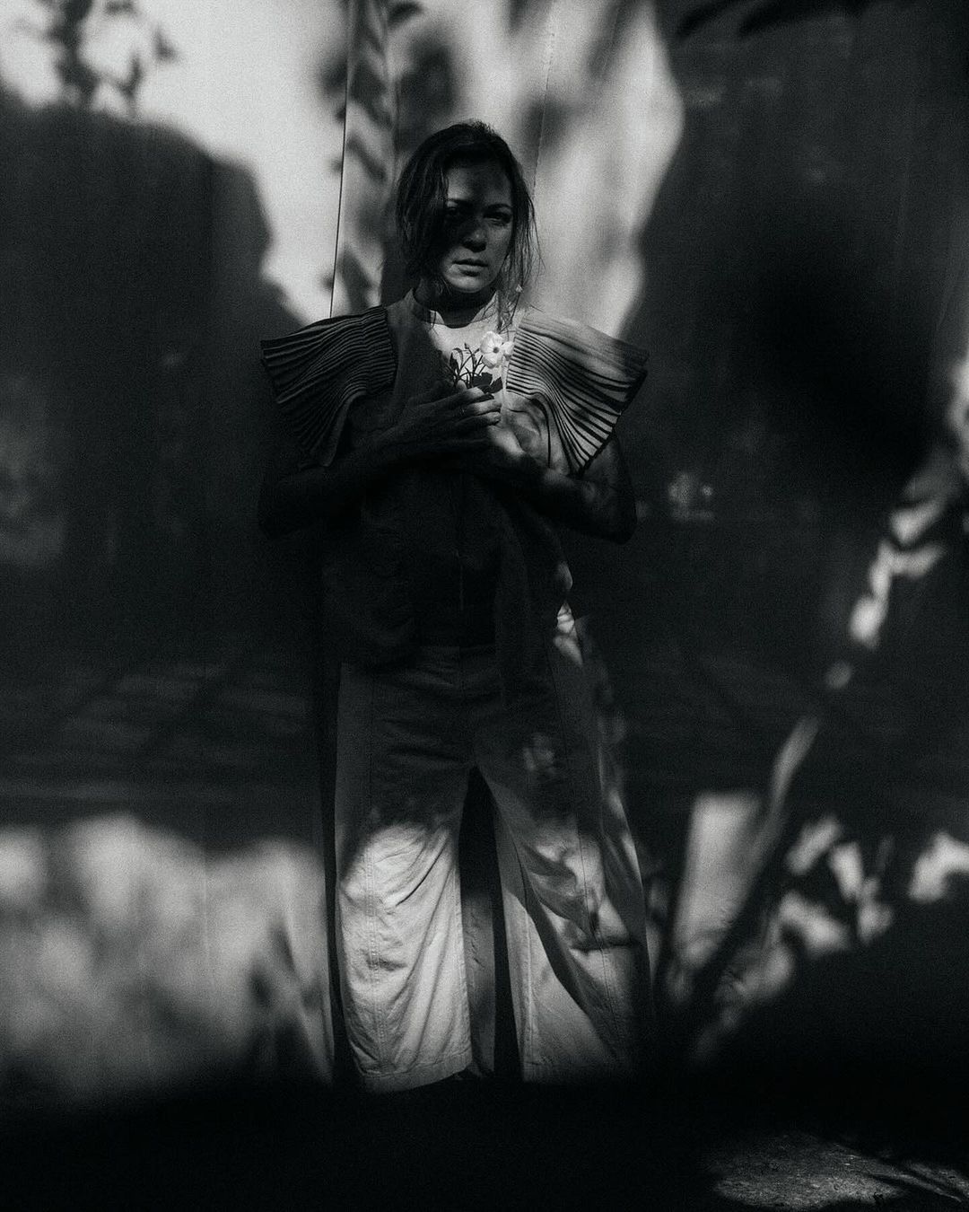 Black and white portrait of a woman in a pleated top holding flowers, standing against a wall with shadows, looking at the camera.