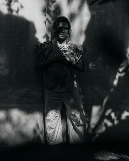 Black and white portrait of a woman in a pleated top holding flowers, standing against a wall with shadows, looking at the camera.