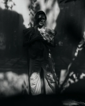 Black and white portrait of a woman in a pleated top holding flowers, standing against a wall with shadows, looking at the camera.