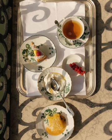 Overhead shot of a silver tray with tea, cake, yogurt, and berries on ornate plates, styled with flowers and shadows.