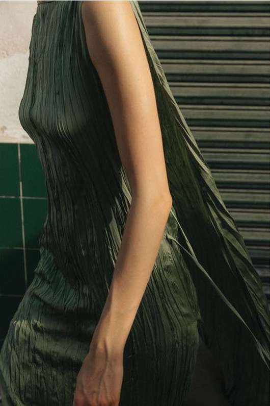 A woman in a pleated, sleeveless dress, with light skin tone, stands against a striped background, bathed in diffused natural light.