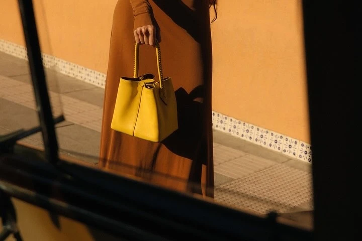 Woman in a brown dress holds a bright yellow handbag outdoors against an orange wall with decorative tile details.