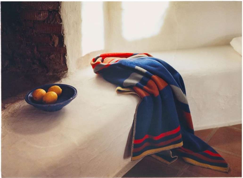 Still life of a striped blanket draped on a bed with a bowl of lemons next to a brick wall in a brightly lit room.