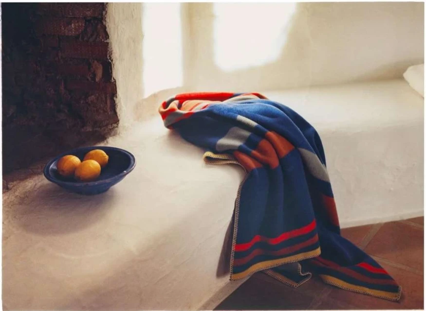 Still life of a striped blanket draped on a bed with a bowl of lemons next to a brick wall in a brightly lit room.