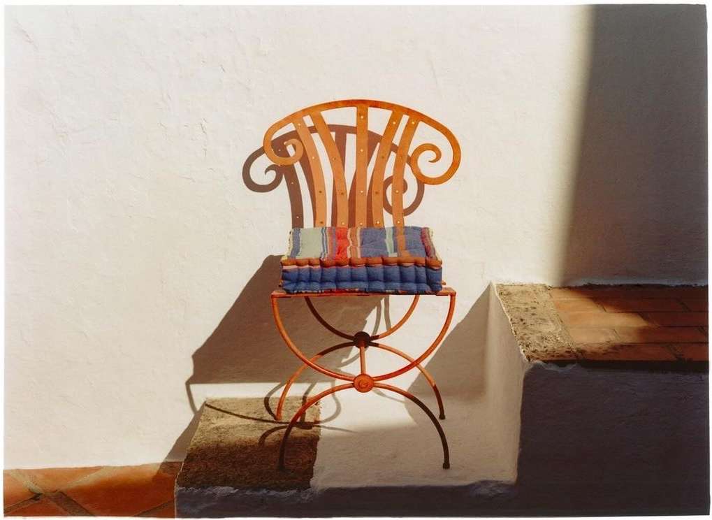 Wrought iron chair with colorful cushion sits on a landing outside a stucco wall, lit by strong sunlight casting dark shadows.