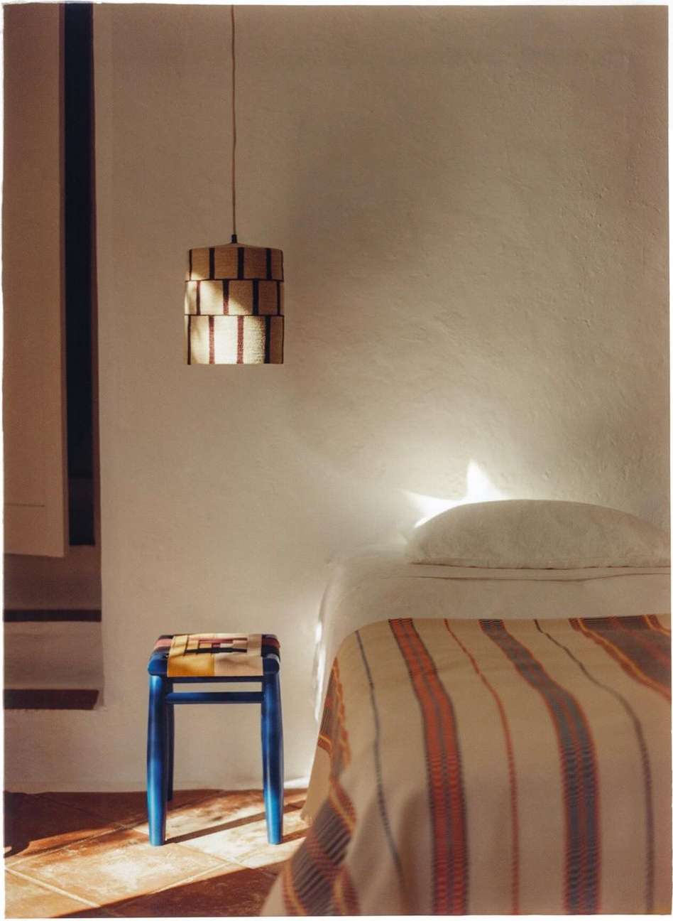 Cozy bedroom scene features a striped blanket on a bed, a blue stool, and a modern pendant light against a white wall.