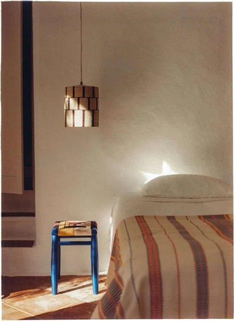 Cozy bedroom scene features a striped blanket on a bed, a blue stool, and a modern pendant light against a white wall.