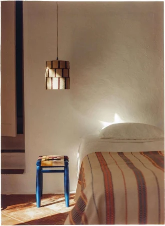 Cozy bedroom scene features a striped blanket on a bed, a blue stool, and a modern pendant light against a white wall.