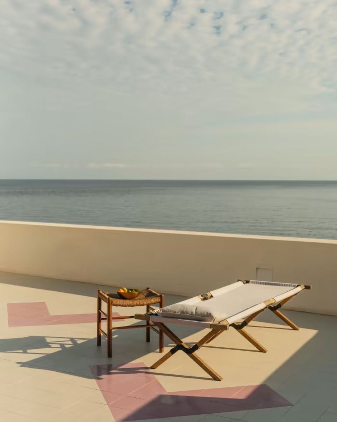 Outdoor lounge area with a daybed and side table on a tiled terrace overlooking the ocean, featuring a serene coastal setting and soft lighting.