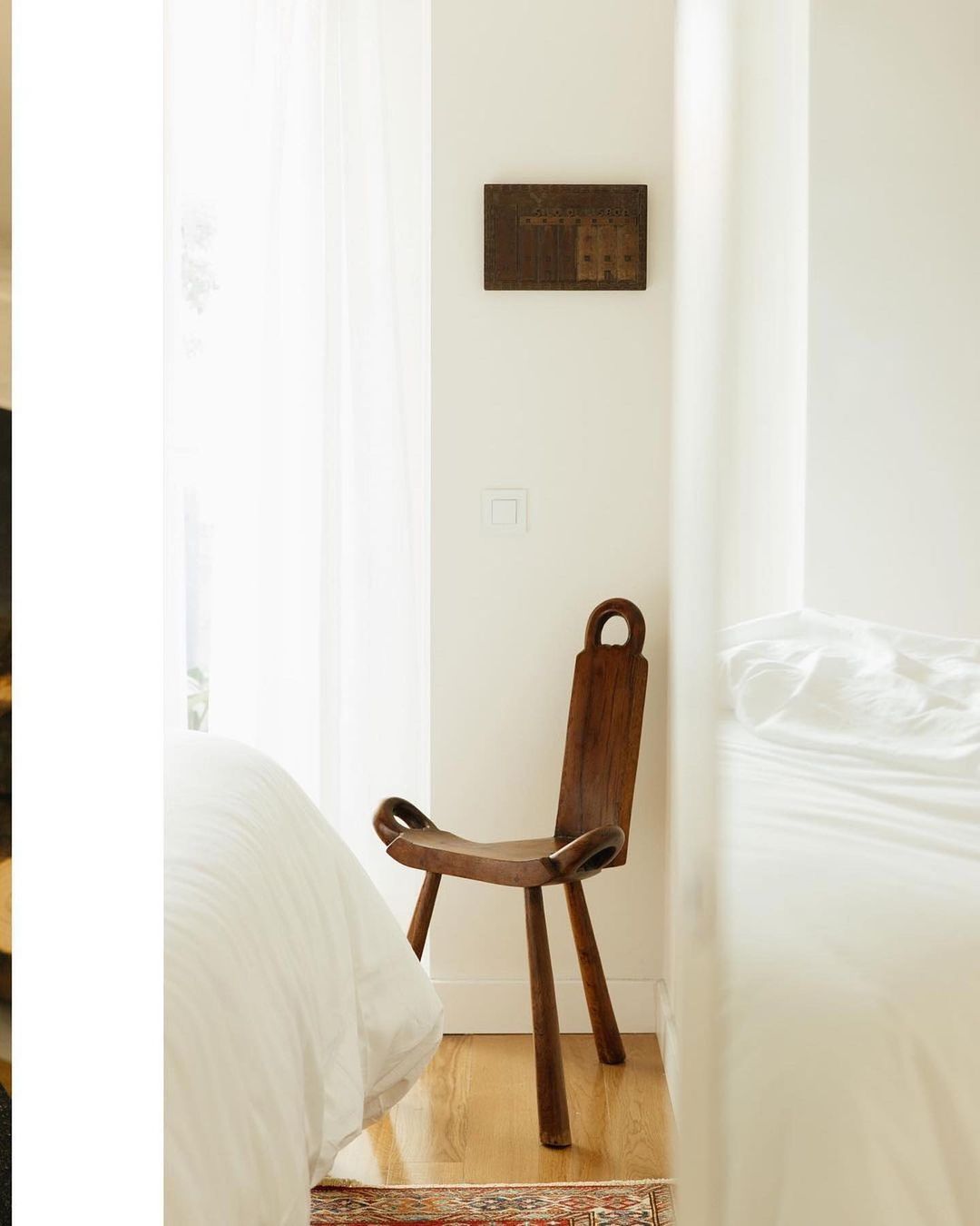 Bedroom corner with a wooden chair and bed featuring white linens. A wooden artwork piece hangs above the chair on the wall.