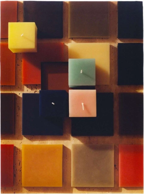 Overhead shot of an array of square candles in various colors including yellow, pink, and blue, arranged on a textured surface.