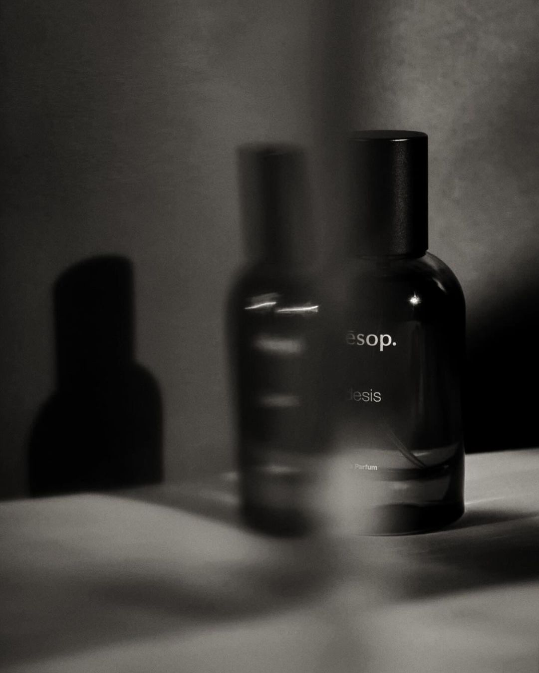 Still life shot of two dark glass Aesop bottles with black caps on a textured white surface in a softly lit grey environment.