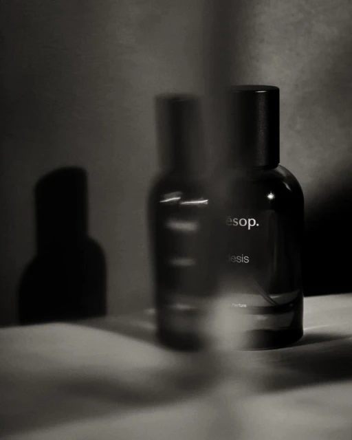 Still life shot of two dark glass Aesop bottles with black caps on a textured white surface in a softly lit grey environment.