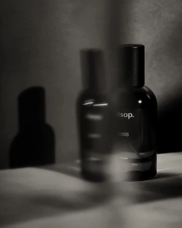 Still life shot of two dark glass Aesop bottles with black caps on a textured white surface in a softly lit grey environment.