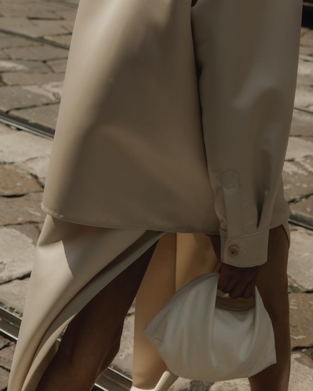 Woman in beige jacket and skirt holds a white clutch. Street background. Fashion editorial image featuring neutral colors.