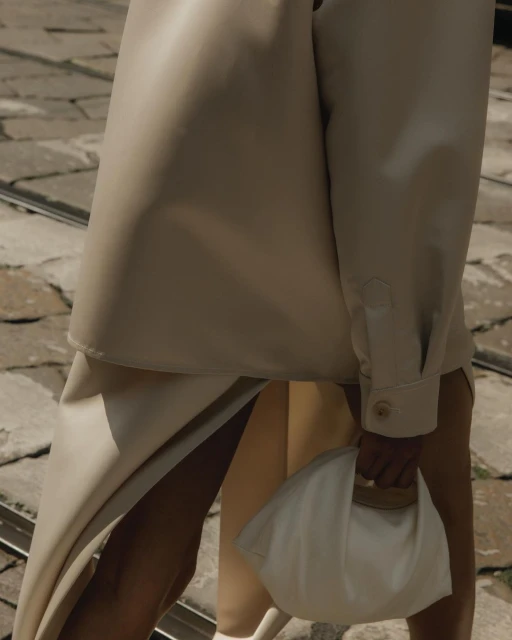 Woman in beige jacket and skirt holds a white clutch. Street background. Fashion editorial image featuring neutral colors.