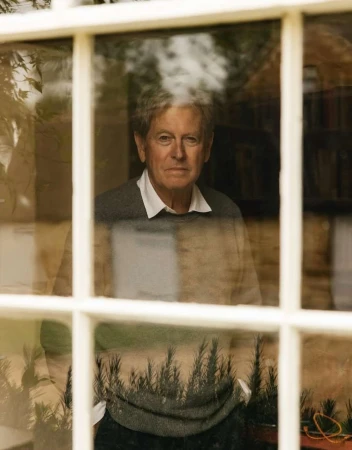 Portrait of an adult man looking through a window; the image reflects plants and foliage outside, creating a multilayered effect.