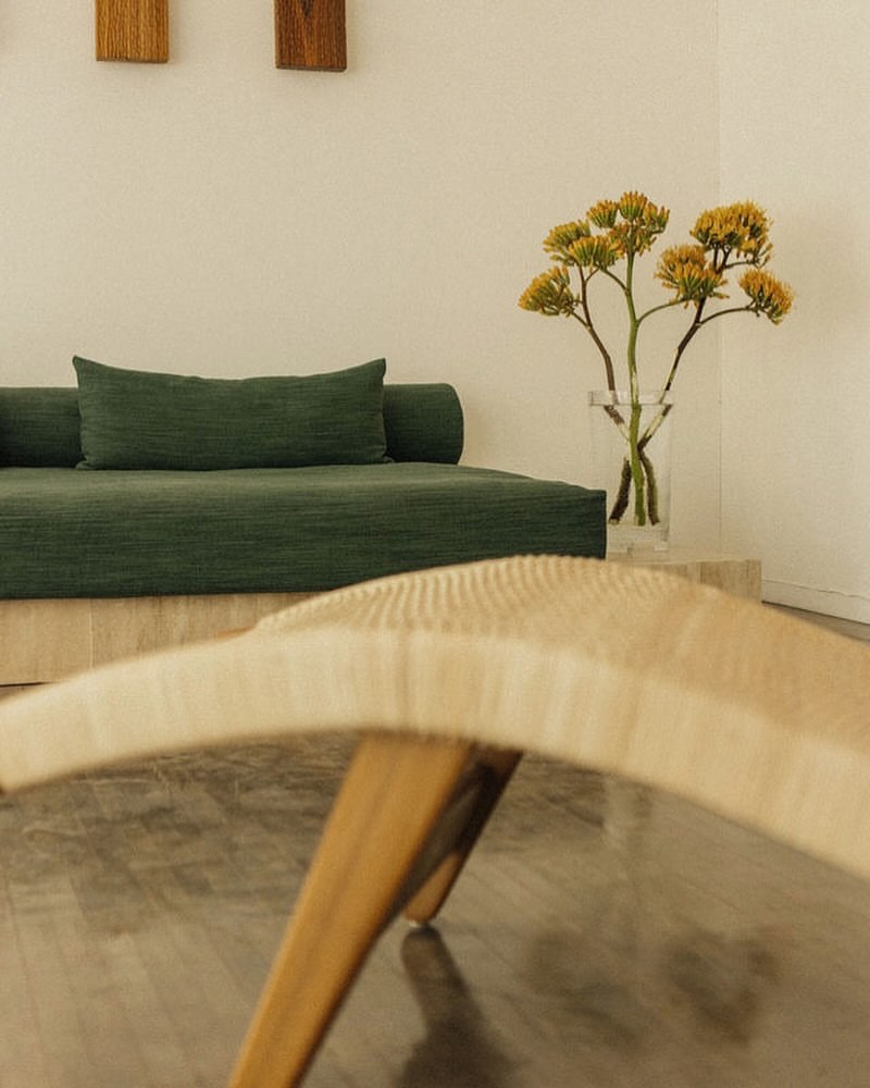 Minimalist interior with a green daybed, clear vase with yellow flowers, and a woven bench, creating a serene and inviting space.