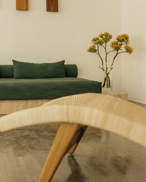 Minimalist interior with a green daybed, clear vase with yellow flowers, and a woven bench, creating a serene and inviting space.