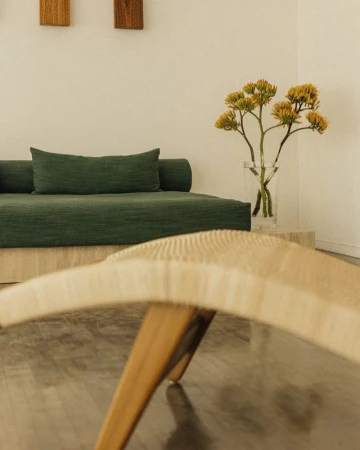 Minimalist interior with a green daybed, clear vase with yellow flowers, and a woven bench, creating a serene and inviting space.