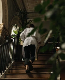 A person seen from behind in a white shirt and black trousers walking up a wooden staircase inside an ornate interior.