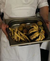Chef in white apron holding a stainless steel tray filled with yellow beans, small round fruits, and a striped squash