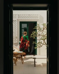 Woman wearing a colourful floral print kaftan walking through a green doorway into a sunlit courtyard in London.