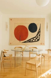 Interior dining room view with a glass table, wooden chairs, and a large abstract wall hanging by Maureen M. Evans