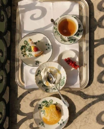 Overhead view of a silver tray with floral-patterned porcelain plates holding tea, cake, yogurt with honey, and red currants in bright sunlight.