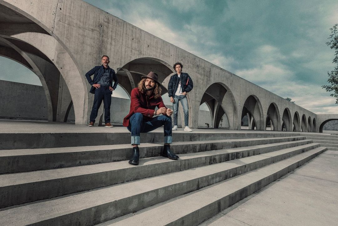 Three men pose on concrete steps in front of an arched building. The man in the center is seated with a hat and red jacket.