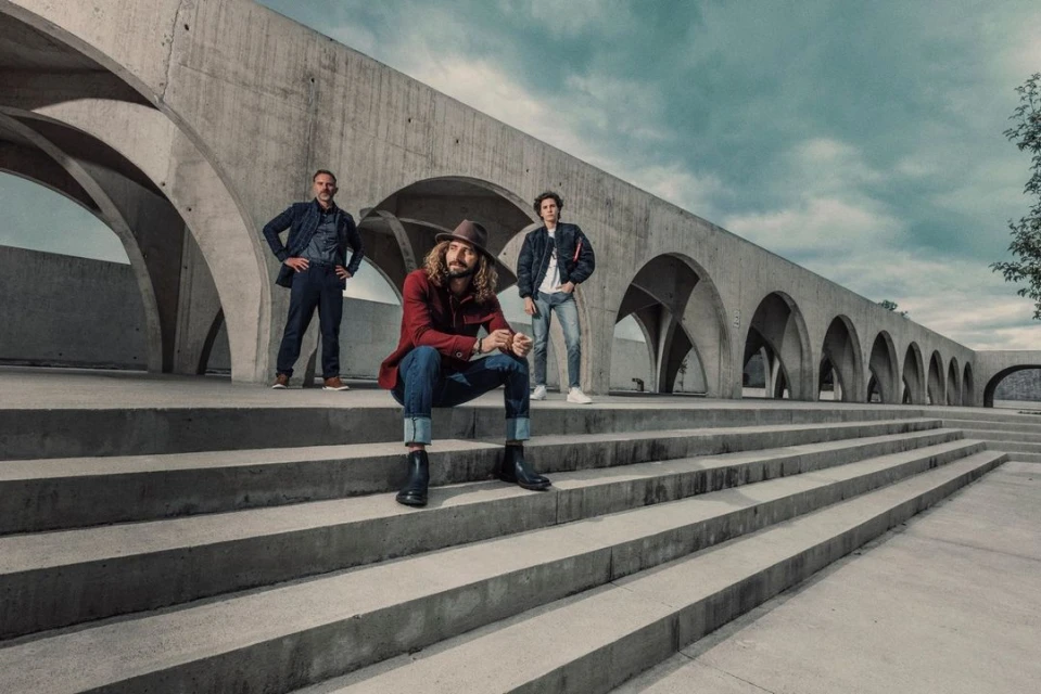 Three men pose on concrete steps in front of an arched building. The man in the center is seated with a hat and red jacket.