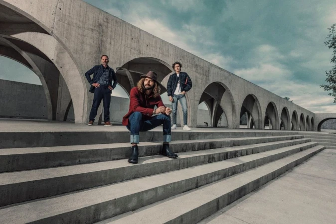 Three men pose on concrete steps in front of an arched building. The man in the center is seated with a hat and red jacket.