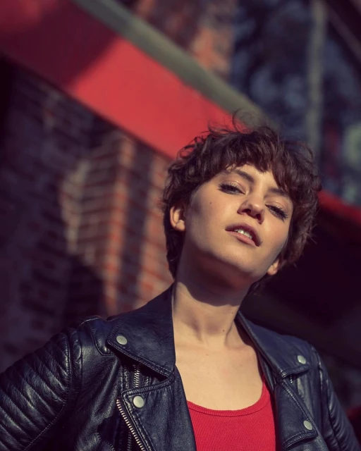 Portrait of a young woman with short brown hair wearing a black leather jacket and red top, posing against a textured brick wall.