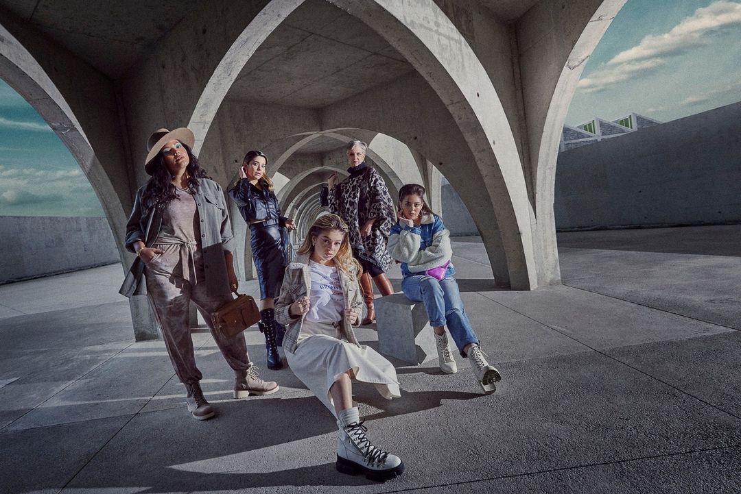 Group of five diverse women posing in fashionable outfits under concrete arches, showcasing modern urban style with neutral tones and textures.