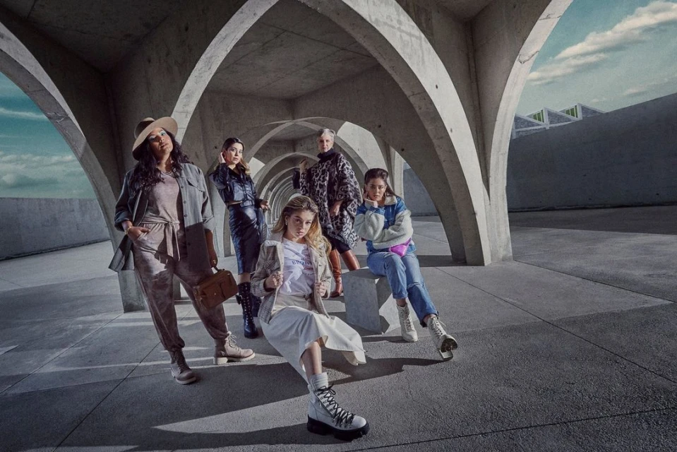 Group of five diverse women posing in fashionable outfits under concrete arches, showcasing modern urban style with neutral tones and textures.