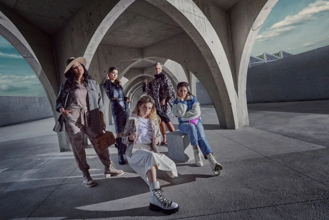 Group of five diverse women posing in fashionable outfits under concrete arches, showcasing modern urban style with neutral tones and textures.