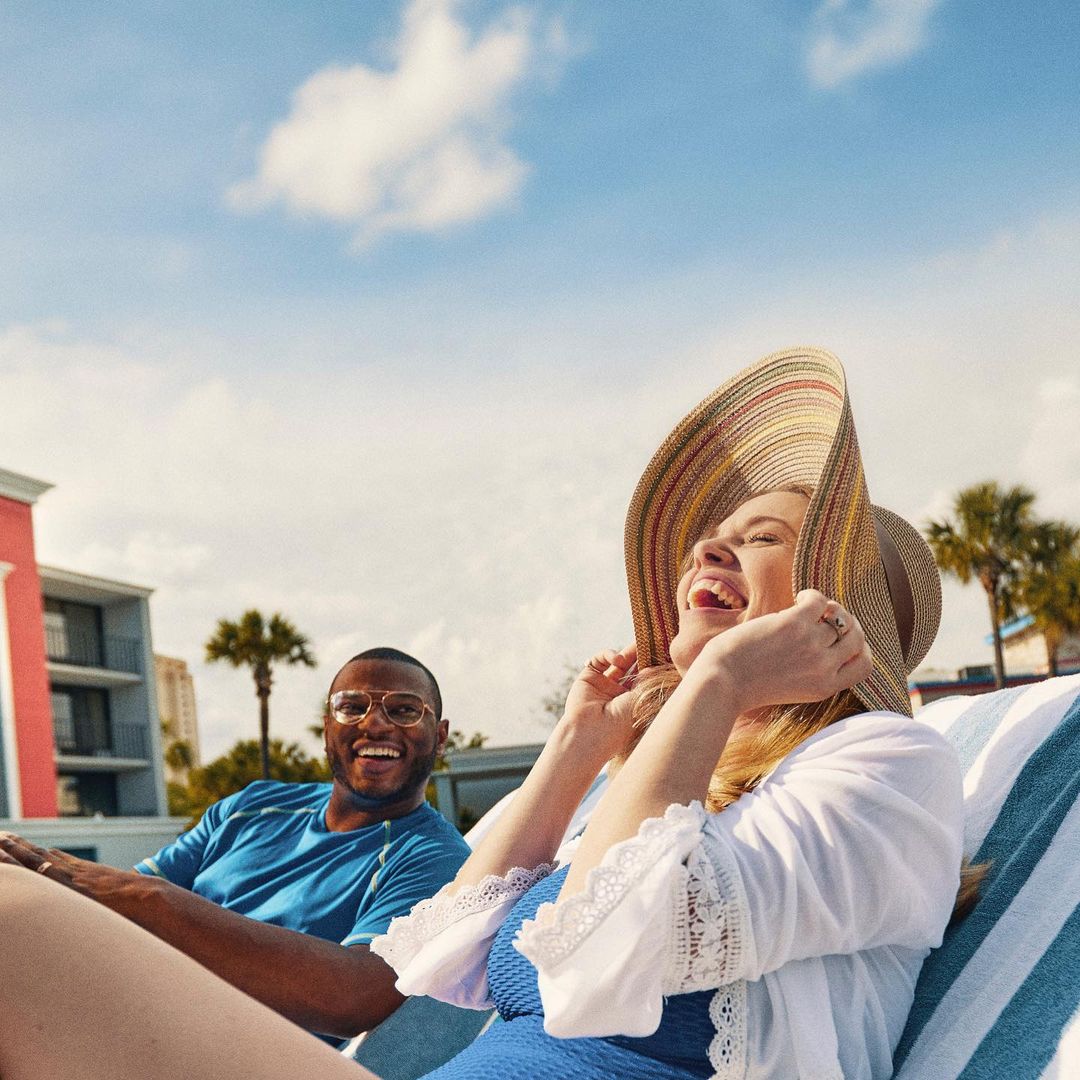 Laughing woman in sunhat relaxes on lounge chair next to smiling man outside on a sunny day with palm trees and blue sky with clouds.