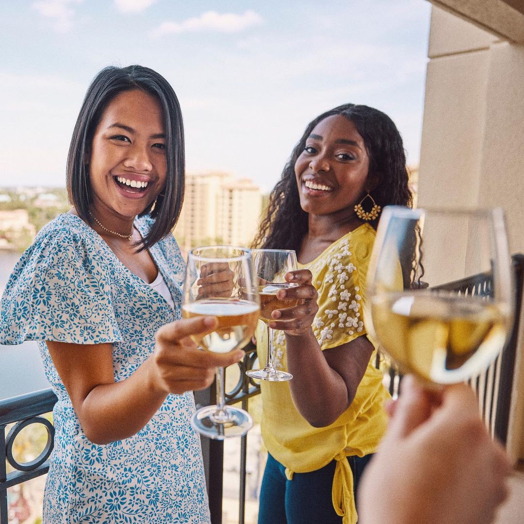 Two smiling women clink wine glasses on a balcony overlooking city buildings and water under a sunny sky, celebrating friendship.