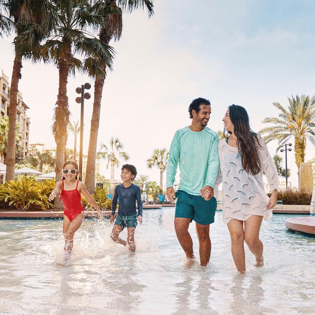 A family of four walks through a shallow swimming pool on a sunny day, surrounded by palm trees and resort buildings.