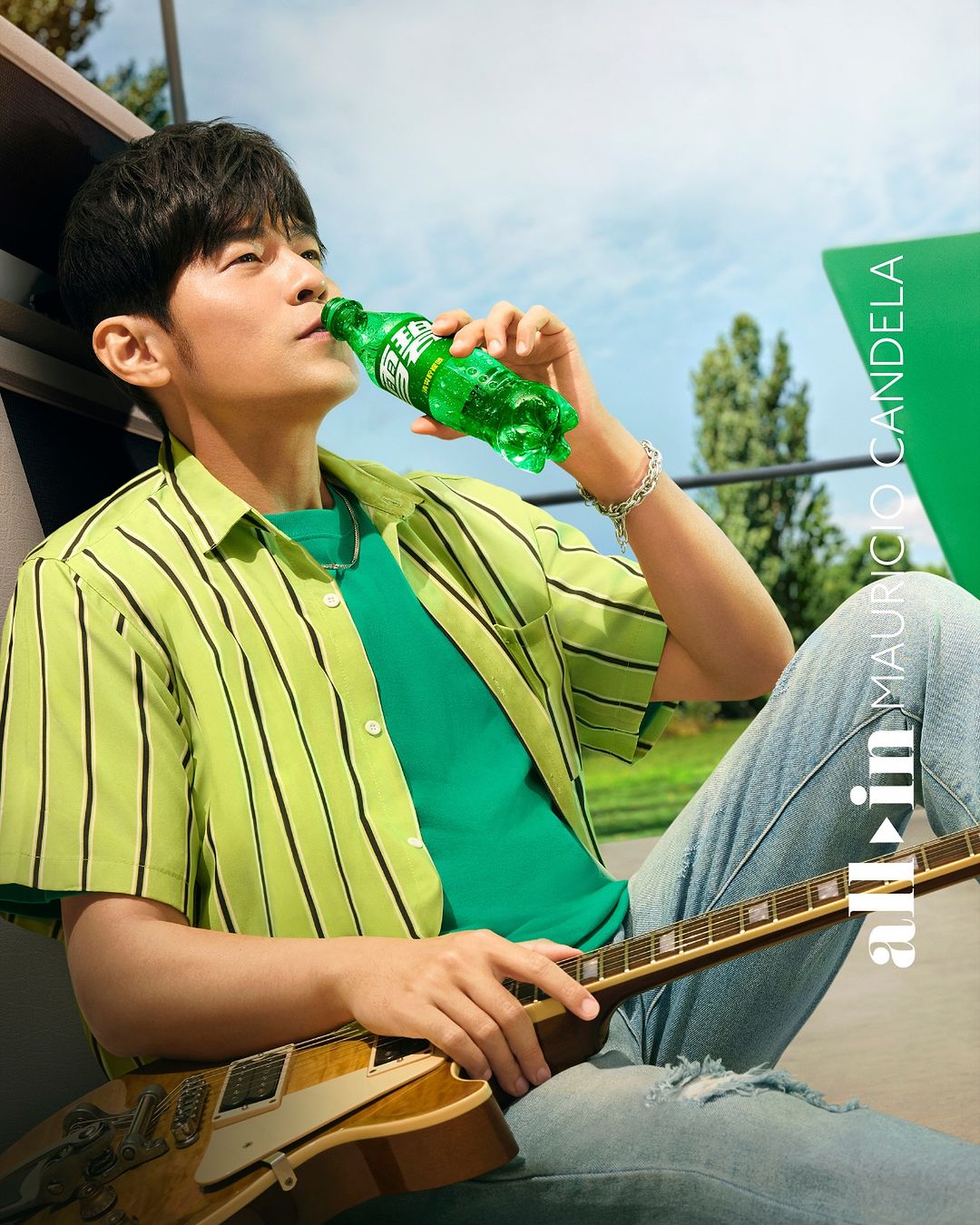Adult male in lime shirt drinking from a green bottle while holding a guitar, with a bright green and blue outdoor background.