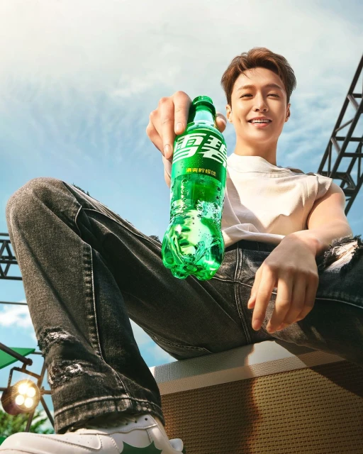 Promotional shot of a smiling man holding a green soda bottle, wearing ripped jeans and sneakers, set against a sky backdrop.